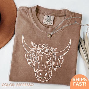 Könnte beinhalten: Espressofarbenes T-Shirt mit weißer Grafik einer Highland-Kuh mit floralen Akzenten. Das Shirt hat einen Rundhalsausschnitt und kurze Ärmel. Das Etikett zeigt "Comfort Colors". Größe S.