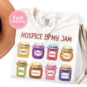 May include: A cream-colored t-shirt featuring the red text "HOSPICE IS MY JAM" above eight colorful illustrations of jam jars. Each jar has a label with words including honor, comfort, dignity, support, person-centered, compassion, quality, and holistic.