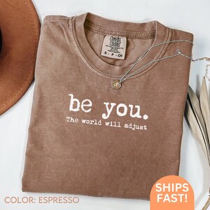 May include: Espresso-colored t-shirt with the text "be you. The world will adjust" in white. The shirt has a crew neck and a Comfort Colors tag. A silver necklace with a pendant is draped across the shirt.