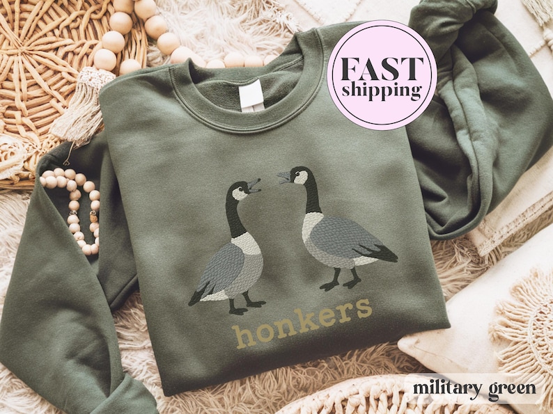 May include: Military green sweatshirt featuring two embroidered geese facing each other. The word "honkers" is embroidered below the geese in a gold color. The sweatshirt is a crew neck style.