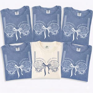 Pode incluir: Seis camisetas são exibidas, cinco em azul e uma em creme. Cada camisa apresenta um design de sutiã de concha com o texto "Margaret's Beach Bach Miami 2016". As camisas estão bem dobradas.