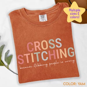 May include: A yam-coloured t-shirt featuring the words "CROSS STITCHING" in pastel shades. The phrase "because stabbing people is wrong" is printed below. The shirt has a Comfort Colors label and is available in multiple sizes and colours.