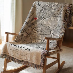 May include: A personalized throw blanket draped over a wooden rocking chair. The blanket features a map design with a red heart and text that reads "ETHAN & SOFIA". Additional text includes coordinates, location, and date. The blanket has fringed edges.