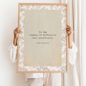 May include: A framed print featuring the quote "In the middle of difficulty lies opportunity" by Albert Einstein. The print has a light brown background with the quote in a typewriter font. The frame is light wood with a floral pattern.