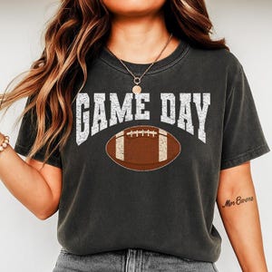 Könnte beinhalten: Dunkelgraues T-Shirt mit dem weißen Schriftzug "GAME DAY" über einer braunen Football-Grafik. Ein lässiges Oberteil, ideal für Sportfans.