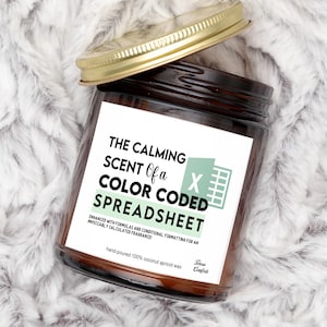 May include: A brown glass jar candle with a gold lid. The label reads "THE CALMING SCENT Of a COLOR CODED SPREADSHEET." The candle is hand-poured with 100% coconut apricot wax. The background is a soft, white, fluffy material.