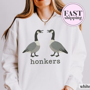 May include: White sweatshirt featuring two embroidered geese facing each other, with the word "honkers" in gray below. The geese are gray and white with black necks. The sweatshirt has a round neck.