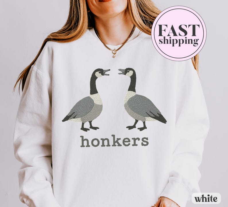 May include: White sweatshirt featuring two embroidered geese facing each other, with the word "honkers" in gray below. The geese are gray and white with black necks. The sweatshirt has a round neck.