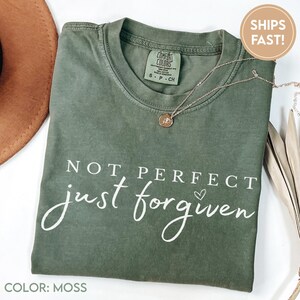 Op de afbeelding: Een mosgroen t-shirt met de tekst "NOT PERFECT just forgiven" in witte letters. Een gouden ketting met hanger ligt op het gevouwen shirt. Het shirt heeft een Comfort Colors label.