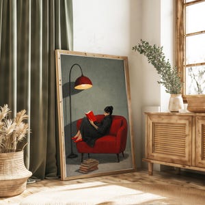 May include: A framed wall art print depicting a person in a black dress and red shoes sitting on a red armchair, reading a red book under a tall black floor lamp. A stack of books rests on the floor beside the chair against a muted green wall.