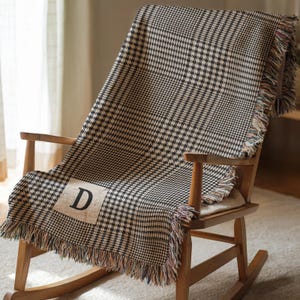 Op de afbeelding: Een zwart-wit geruite plaid met franjes en de letter "D" op een patch. De plaid is over een houten schommelstoel gedrapeerd, wat een gezellige en uitnodigende scène creëert. De franjes zijn veelkleurig.