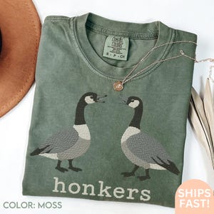 May include: Moss green t-shirt featuring two geese facing each other, with the word "honkers" printed below. The shirt has a crew neck and short sleeves. A gold necklace is draped across the shirt. The shirt is made of a soft, comfortable material.