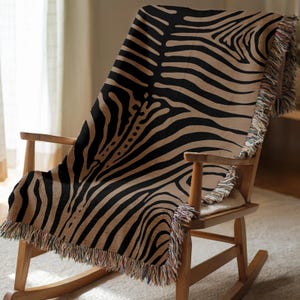 May include: A tan and black zebra print throw blanket draped over a wooden rocking chair. The blanket has a fringed edge. The chair is made of light-coloured wood. The blanket is made of a soft material.