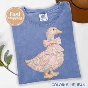 Pode incluir: Camiseta azul jeans com uma aplicação de pato em patchwork. O pato apresenta um laço rosa e tecidos estampados com flores. A camiseta tem gola redonda e mangas curtas.