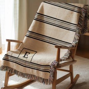 Op de afbeelding: Een beige sprei met zwarte horizontale strepen en franjes is over een houten schommelstoel gedrapeerd. De sprei heeft de letter "E" in een zwart vierkant. Een gezellig woondecoratie-item.