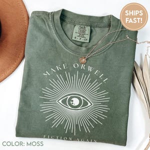 May include: Moss green t-shirt with the text "MAKE ORWELL FICTION AGAIN" and an eye graphic. A gold necklace with a pendant rests on the shirt. The shirt is folded and next to a brown hat.
