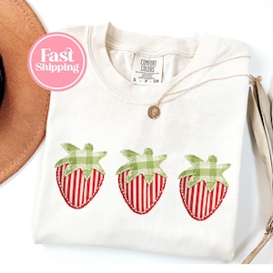 May include: Cream-coloured t-shirt featuring three strawberry appliqués. Each strawberry has red and white striped detailing and green gingham tops. A gold necklace with a pendant rests on the shirt. The shirt has a Comfort Colors tag.