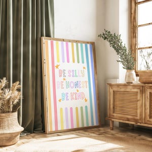 Op de afbeelding: Een ingelijste kunstdruk met een houten frame. Het kunstwerk toont verticale strepen in pastelkleuren en de woorden "BE SILLY, BE HONEST, BE KIND" in kleurrijke tekst. Gouden sterren accentueren de tekst. De print wordt tentoongesteld in een kamer met natuurlijk licht.