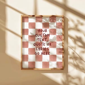 Op de afbeelding: Een ingelijste print met een roze en wit geblokt patroon. De print bevat de tekst "YOUR CUSTOM TEXT, QUOTE OR LYRICS GO HERE" in een handschriftstijl. Het frame is van hout.