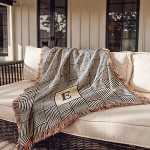 Op de afbeelding: Een geweven sprei met een geometrisch patroon in zwart en beige, gedrapeerd over een beige buitenzitbank. De deken heeft een grote letter "E" in het midden en gefranjerde randen. De bank heeft witte kussens.