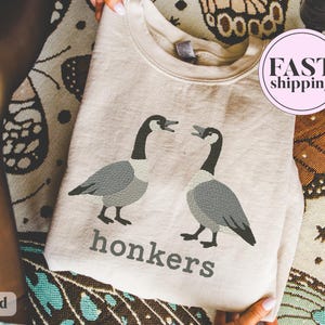 May include: Sand-colored sweatshirt featuring a graphic of two geese with open beaks, in shades of gray and black. Below the geese, the word "honkers" is printed in gray. A pink circle with "FAST shipping" is visible.