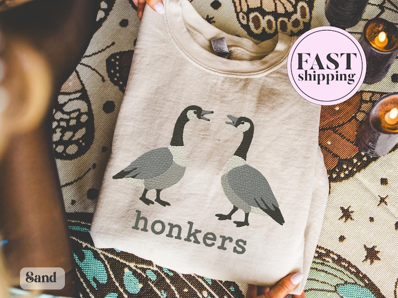 May include: Sand-colored sweatshirt featuring a graphic of two geese with open beaks, in shades of gray and black. Below the geese, the word "honkers" is printed in gray. A pink circle with "FAST shipping" is visible.