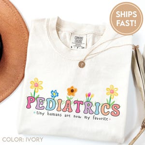 May include: Ivory t-shirt with the word "PEDIATRICS" in colourful, bubble letters, and the phrase "tiny humans are now my favourite." The shirt features a floral design and a gold necklace. The shirt is made by Comfort Colors.