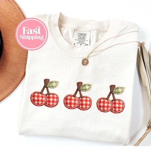 Puede incluir: Camiseta de color crema con tres diseños de cerezas bordadas. Cada cereza es de cuadros rojos y blancos con tallos marrones y hojas verdes. Un collar dorado con un pequeño colgante descansa sobre la camiseta. La camiseta tiene una etiqueta "Comfort Colors".