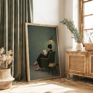 May include: A framed rectangular wall art print depicting a person in a dark dress sitting in a green armchair, reading a book under a floor lamp. The scene includes a small side table with stacked books, all set against a muted green wall.