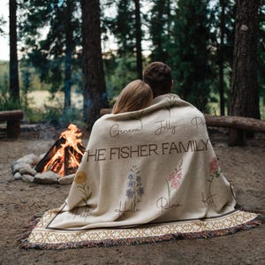 May include: Cream-coloured throw blanket with floral embroidery and the text "THE FISHER FAMILY". The blanket has a decorative border and fringe. A couple is sitting near a campfire, wrapped in the blanket.