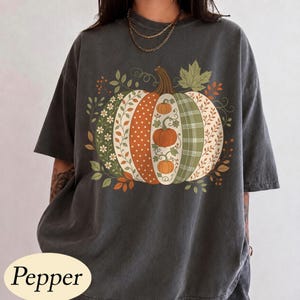以下が含まれることがあります： チャコールグレーの半袖Tシャツ。中央には、水玉模様、チェック柄、花のつるなど、様々なパターンで垂直に分割されたカボチャの大きなグラフィックが描かれており、小さなオレンジ色のカボチャと葉がアクセントになっています。