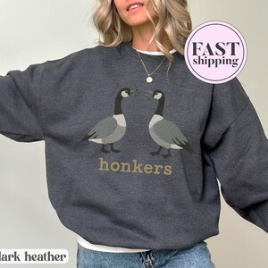 May include: Dark heather gray sweatshirt featuring two geese facing each other, with the word "honkers" in gold lettering below. The sweatshirt has a crew neck and long sleeves. The words "dark heather" are printed at the bottom.