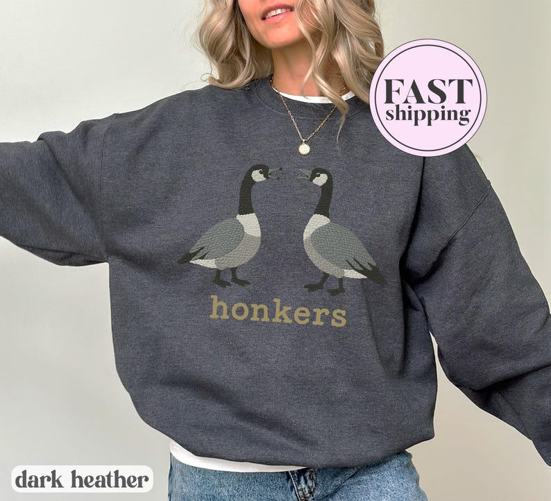May include: Dark heather gray sweatshirt featuring two geese facing each other, with the word "honkers" in gold lettering below. The sweatshirt has a crew neck and long sleeves. The words "dark heather" are printed at the bottom.