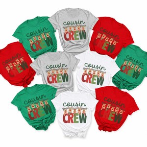 May include: A collection of short-sleeved t-shirts in red, green, white, and gray. Each shirt features the text "cousin CREW" in a dark green and red font, accompanied by a graphic of five reindeer wearing colorful scarves hanging from the letters.