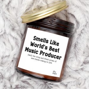 May include: A brown glass candle jar with a gold lid, featuring a white label with the text "Smells Like World's Best Music Producer." The label also includes the text "Smell the crisp and fresh aroma of new tracks coming to life."