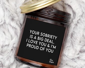 Puede incluir: Una vela en tarro de cristal marrón con tapa dorada, sobre una superficie blanca y esponjosa. La vela tiene una etiqueta negra con texto blanco: "YOUR SOBRIETY IS A BIG DEAL. I LOVE YOU & I'M PROUD OF YOU."
