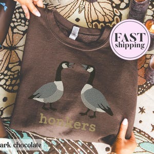 May include: Dark chocolate-colored sweatshirt featuring a graphic of two geese facing each other. The word "honkers" is printed below the geese in a light green color. The sweatshirt is being held up by a person.