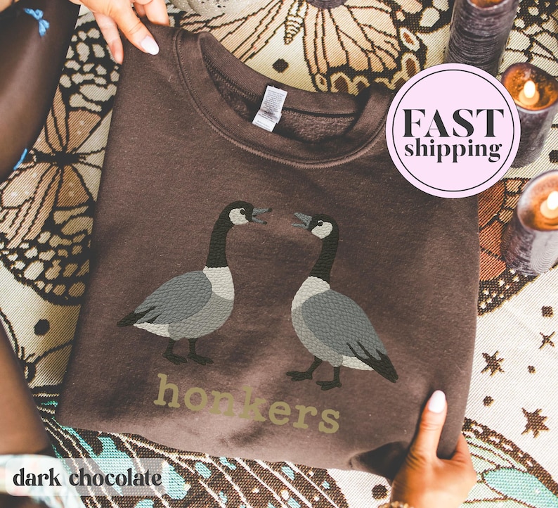 May include: Dark chocolate-colored sweatshirt featuring a graphic of two geese facing each other. The word "honkers" is printed below the geese in a light green color. The sweatshirt is being held up by a person.