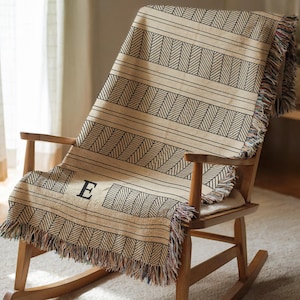 Op de afbeelding: Een beige sprei met een zwart geometrisch patroon en franjes is over een houten schommelstoel gedrapeerd. De sprei heeft een herhalend ontwerp van diagonale lijnen en horizontale strepen. De letter "E" is op de sprei geborduurd.