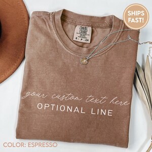 May include: Espresso-colored t-shirt with the text "your custom text here OPTIONAL LINE". The shirt has a crew neck and a silver necklace with a pendant. The shirt is labeled "Comfort Colors" and is size S-CH.