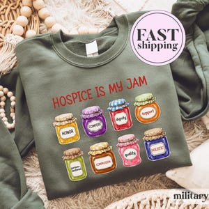 May include: A military green sweatshirt featuring the red text "HOSPICE IS MY JAM" above eight colorful illustrations of glass jars. Each jar is labeled with words including honor, comfort, dignity, support, person-centered, compassion, quality, and holistic.