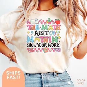 Peut inclure: T-shirt ivoire avec un motif coloré indiquant "THE MATH AIN'T MATHIN SHOW YOUR WORK". Le design comprend une pomme, des ciseaux, un globe et d'autres éléments scolaires. Le t-shirt est de style décontracté.
