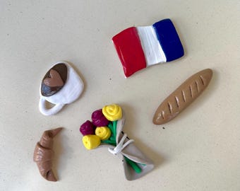 Puede incluir: Colección de miniaturas de comida y objetos con temática francesa. Incluye una taza de café blanca con un corazón, una bandera francesa, una baguette, un cruasán y un ramo de flores amarillas y moradas.