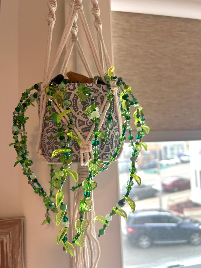 May include: A hanging macrame plant holder with a patterned ceramic pot. The pot is adorned with a beaded garland featuring green beads and leaf accents. The plant holder is suspended by white rope.