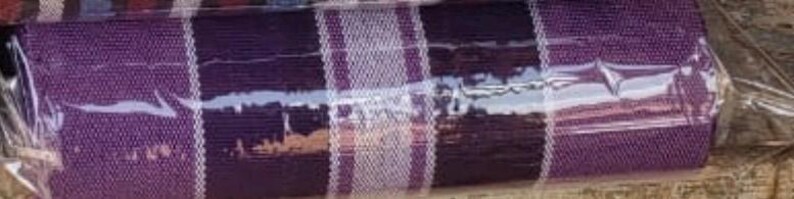 Puede incluir: Una tela textil enrollada apretadamente que presenta un color base morado oscuro con rayas verticales prominentes en tonos blancos y morados m&aacute;s claros. La tela tiene una textura tejida visible y est&aacute; envuelta en un pl&aacute;stico protector transparente.