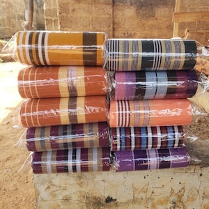 Puede incluir: Una pila de diez rollos de tela tejida rectangulares, cada uno envuelto individualmente en pl&aacute;stico transparente. Los rollos presentan varios patrones de rayas en colores que incluyen amarillo mostaza, naranja, negro, morado, azul y melocot&oacute;n.