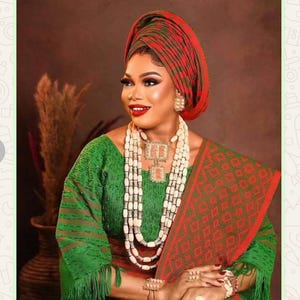 May include: A person wearing a green and red patterned outfit, including a head wrap and shawl. The ensemble is complemented by layered white beaded necklaces, earrings, and bracelets. The outfit has a traditional aesthetic.