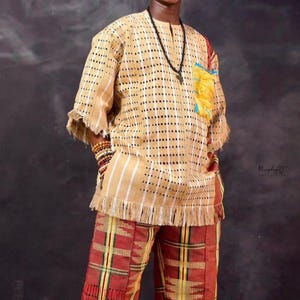 Può includere: Una persona che indossa un abito tradizionale africano. L'ensemble comprende una camicia fantasia beige e bianca con frange, pantaloni abbinati a quadri rossi, gialli e verdi e un berretto giallo. Una collana di perline nere e braccialetti completano il look.