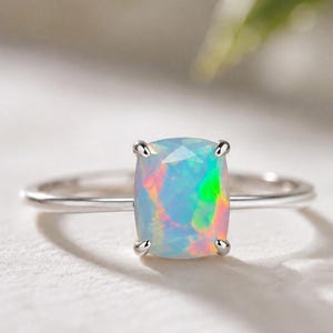 Könnte beinhalten: Ein Silberring mit einem im Kissenschliff facettierten Opal, der von vier Krappen gehalten wird. Der Opal zeigt irisierende Lichtreflexe in Blau, Grün und Pink. Die Ringschiene ist dünn, poliert und metallisch und bildet einen Kontrast.