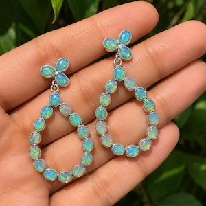 Könnte beinhalten: Ein Paar Ohrhänger mit ovalen, schillernden Opalen in silberfarbenen Fassungen. Jeder Ohrring besteht aus einem tropfenförmigen Rahmen aus mehreren kleinen Opal-Cabochons, der von einem dreiteiligen, kleeblattartigen Element gekrönt wird.